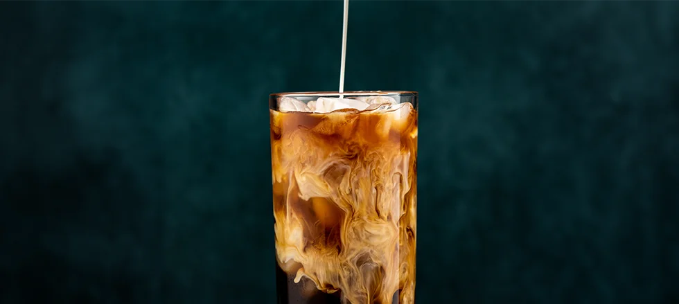 The image features a tall glass filled with a brown beverage, which is likely iced coffee. The glass is sitting on a table, possibly in a restaurant setting.