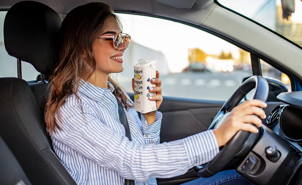 The image features a woman sitting in the driver's seat of a car, holding a cup in her hand. She is wearing a striped shirt and has her car's steering wheel in front of her. The scene suggests that she is driving the car while enjoying her drink.