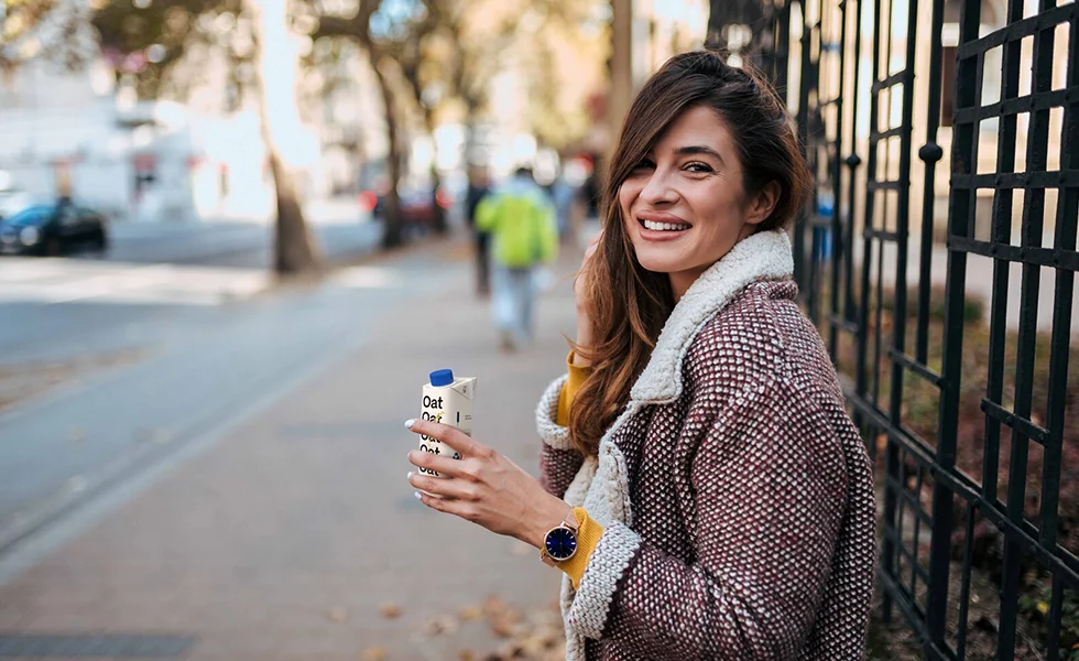 The image features a woman standing on the sidewalk, holding a SIG packaging in her hand. She is wearing a jacket and appears to be smiling.