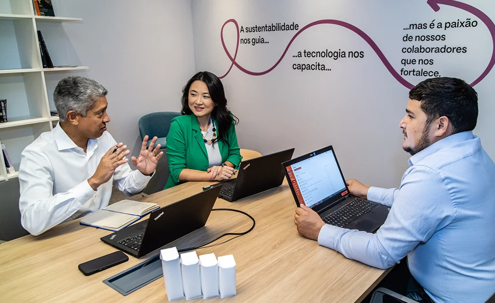 The image features a group of three people sitting around a table, engaged in a discussion or meeting. They are all using laptops, which are placed on the table in front of them. The individuals appear to be professionals, possibly business people, as they are talking and working together.