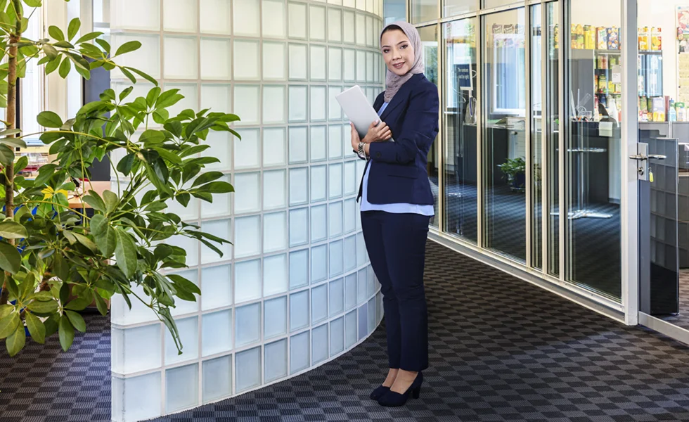 The image features a woman standing in an office area, holding a clipboard or a piece of paper. She is positioned next to a large wall covered with clear, block-like, frosted glass. The wall appears to be a divider in the office, and the woman is standing close to it. The office setting suggests that the woman might be an employee or a manager, possibly reviewing some documents or preparing for a meeting.