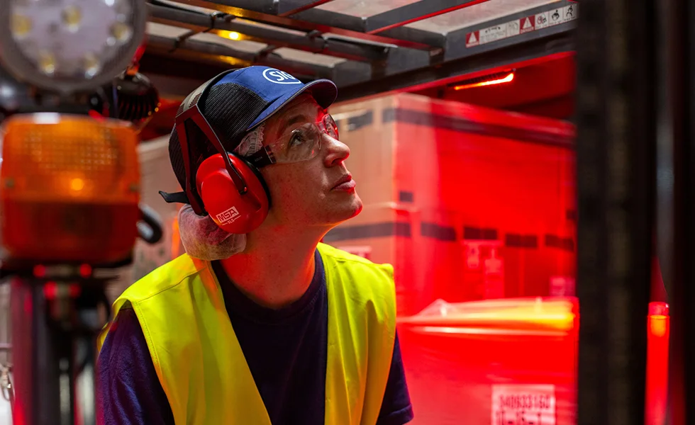 The image features a person wearing safety goggles and a yellow vest, standing in front of a red curtain. She or he appears to be looking at something, possibly a display or an event happening behind the curtain. 