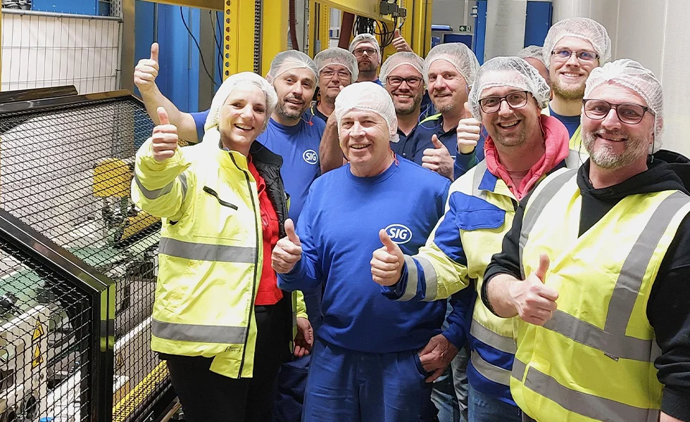 The image features a group of workers, possibly construction workers, posing for a photo. They are wearing hard hats and are gathered together, likely for a group picture. There are at least 10 people in the scene, with some standing closer to the camera and others further back. The workers are giving thumbs up, indicating their satisfaction or approval of the project they are working on.