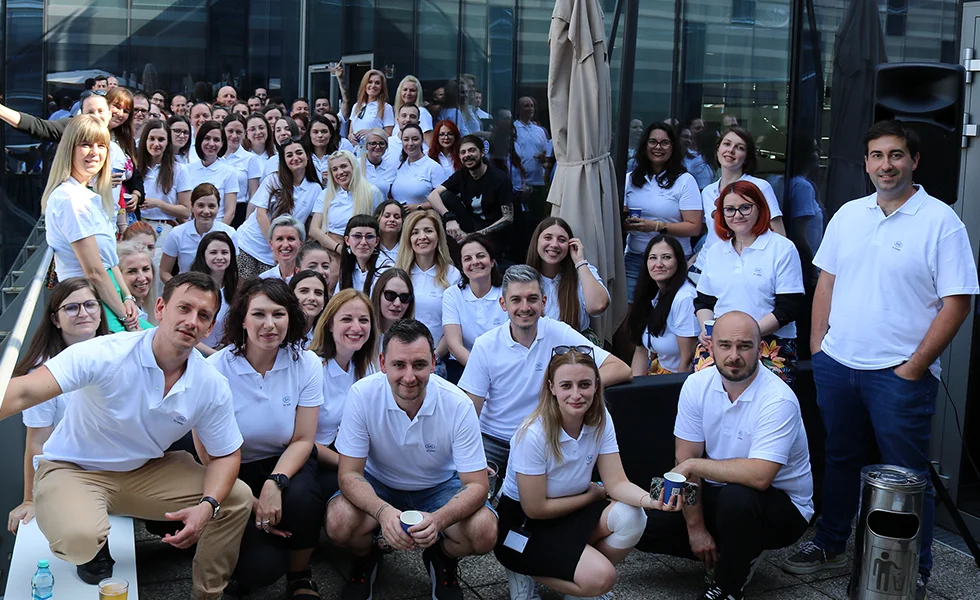 The image features a large group of people, possibly employees, posing for a group photo. They are all wearing white shirts, which indicates they may be part of a team or organization. Some of the people are standing, while others are sitting, and they are arranged in a way that fills the frame of the photo.