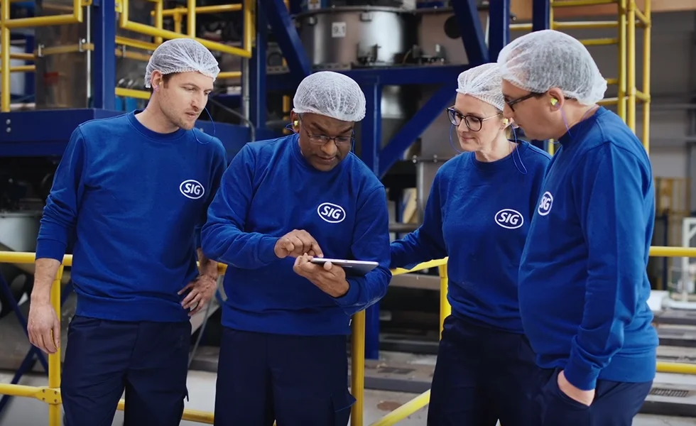 The image features a group of four people, two men and two women, all wearing blue shirts and white hairnets, standing in a factory or warehouse setting. They are looking at a tablet computer, with three of them focusing on the screen while the fourth person, a woman, looks on. The group appears to be engaged in a work-related discussion or task.