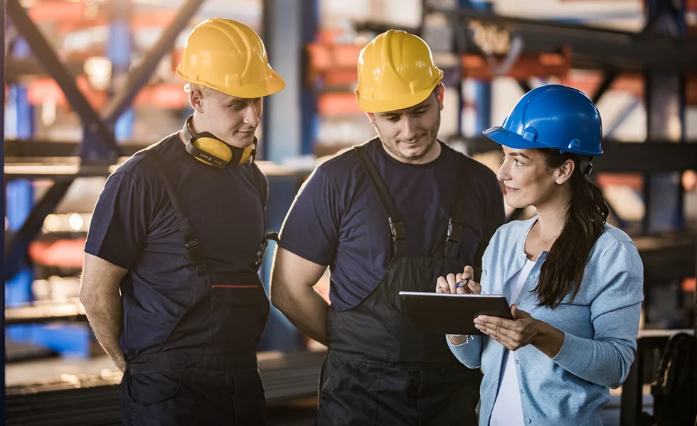 The image features three people in a factory setting, with two men wearing yellow hard hats and a woman in a blue shirt. The woman is holding a tablet and appears to be showing something on the screen to the two workers. They are all standing near a bench.