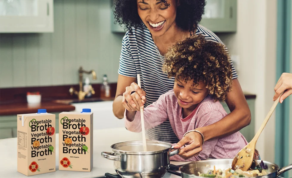 The image features a woman and a child in a kitchen, preparing food together. The woman is showing the child how to make broth. There are several bottles, cups, and a bowl placed around the kitchen, indicating that they are using various ingredients in the process. A spoon is also visible, being used in the preparation. The scene conveys a sense of bonding and learning between the woman and the child as they cook together.