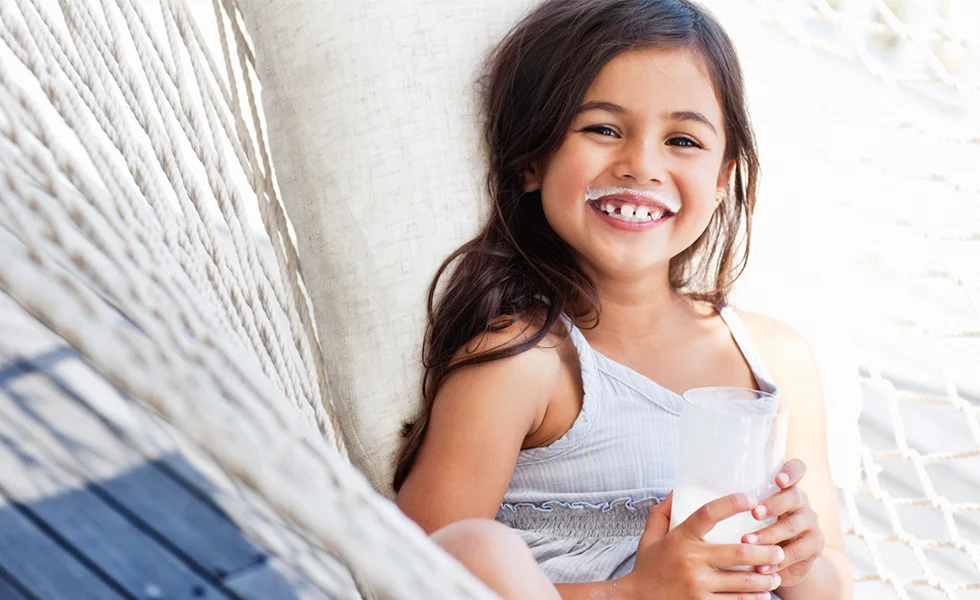 The image features a young girl, likely a toddler, sitting on a hammock while holding a cup of milk in her hand. She is smiling, possibly enjoying her time on the hammock.