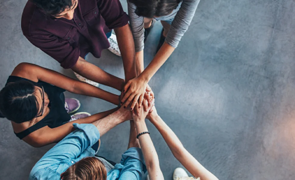 The image features a group of people, likely young adults, standing in a circle and holding hands or giving each other high fives. They appear to be celebrating or showing support for one another.