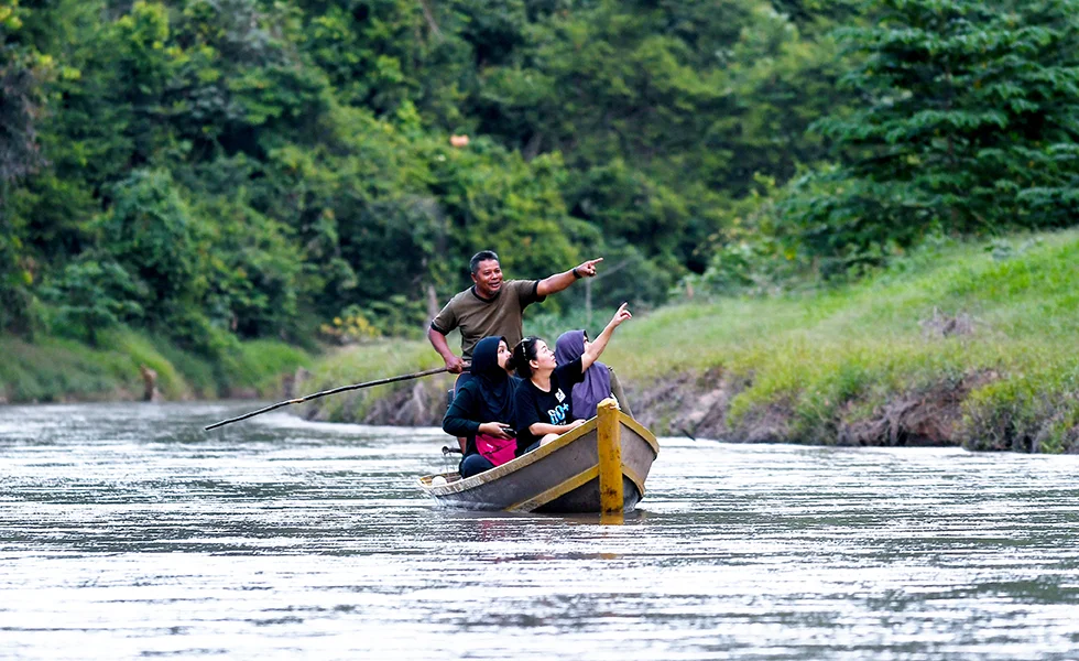 The image features a group of people riding on a small boat, possibly a canoe or a raft, in a river. There are at least five people in the boat, with one person in the front, two in the middle, and two more towards the back. They seem to be enjoying their time together, floating down the river.