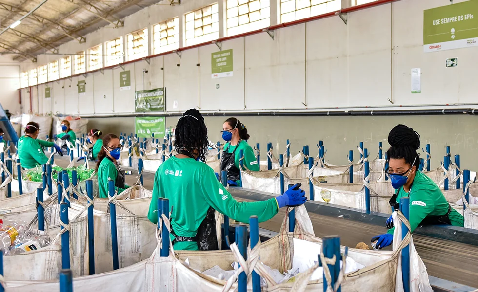 The image shows a group of people, likely workers, wearing green shirts and sorting items, in what appears to be an assembly line setting. They are working in a building with white walls.