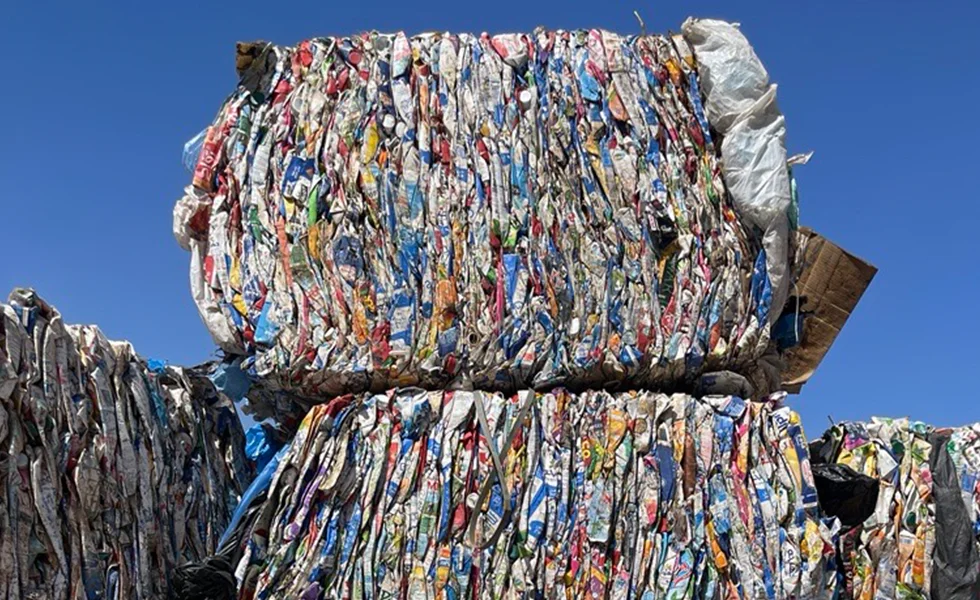 The image features a large pile of recyclable materials, primarily consisting of flattened aluminum cans and plastic bottles. The pile is arranged in a stack, with the cans and bottles in various colors and sizes.