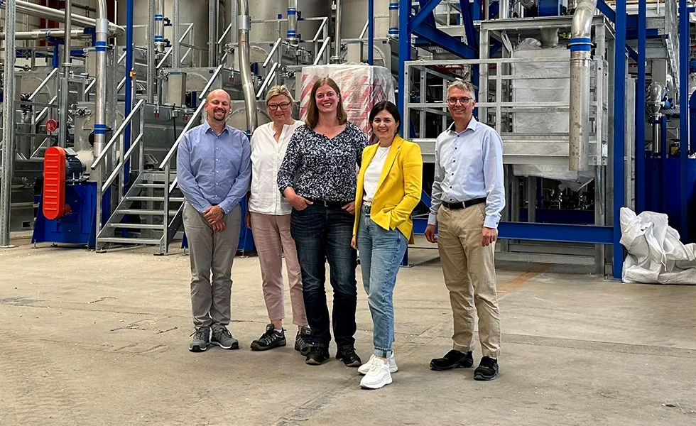 The image features a group of people standing together in a factory or warehouse setting. 