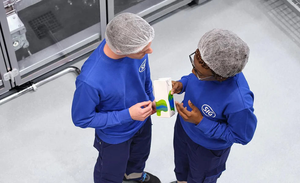 The image features two workers wearing blue and white uniforms, standing next to each other in a factory or work environment. They appear to be having a conversation, possibly discussing work-related matters or sharing personal stories. Both workers are wearing protective gear, such as hairnets, which suggests that they are engaged in some kind of manufacturing or production process.