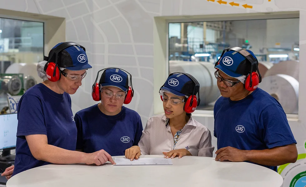 The image features a group of four people, possibly workers, standing around a table and looking at a paper or document together. They are wearing ear protection, likely to protect their hearing in a noisy environment.