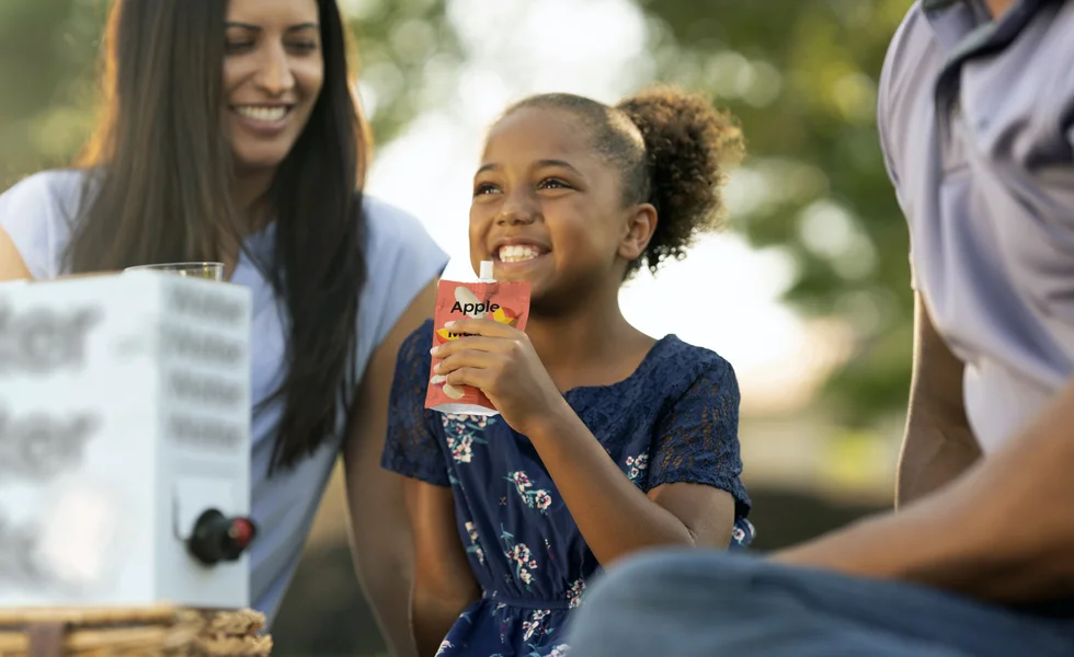 The image features a young girl holding a juice pouch in her hand. She is smiling and enjoying the moment.