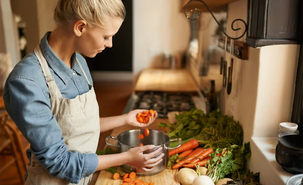 The image features a woman in an apron standing in a kitchen, preparing food. She is holding a bowl and is surrounded by various vegetables, including carrots, which are displayed on the counter. There are also other bowls and bottles in the kitchen, indicating that the woman is actively engaged in cooking or preparing a meal.