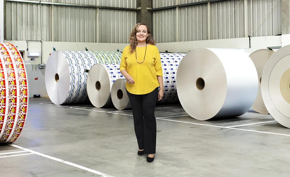 The image features a woman standing in a warehouse or factory setting, surrounded by large rolls. She is posing in front of these rolls, which are stacked on top of each other in various arrangements. The woman appears to be the main subject of the photo, with the rolls serving as a background element that provides context to the scene.